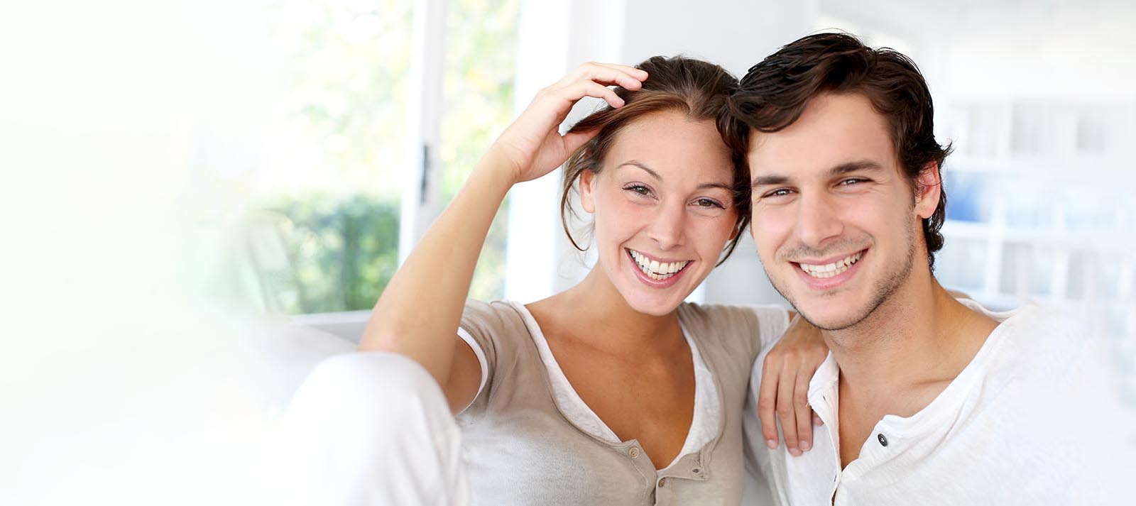 The image shows a man and woman smiling at the camera, with the man standing behind the woman. They appear to be posing for a portrait or photograph together.